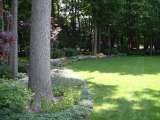 Landscaping design in woodland area has beds with plants and flowers behind backyard lawn.